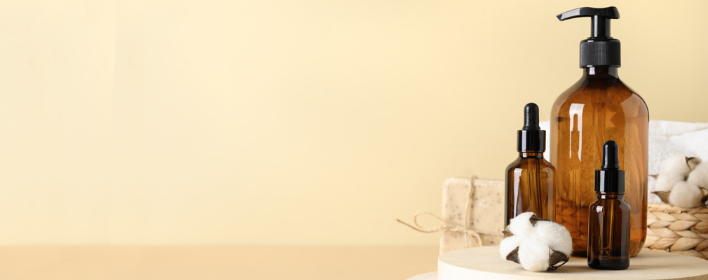 Guida al packaging cosmetico sostenibile Immagine d'atmosfera "imballaggio cosmetico sostenibile": flaconi marroni con pipetta e pompa disposti con cotone, sapone e un cestino di sisal su uno sfondo color albicocca