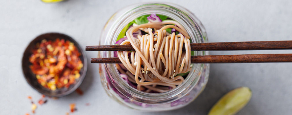 Guida alla preparazione dei pasti Immagine con l'atmosfera "Meal Prep": noodles asiatici in un bicchiere sollevati da due bacchette di legno marrone, accanto a una ciotola di spezie