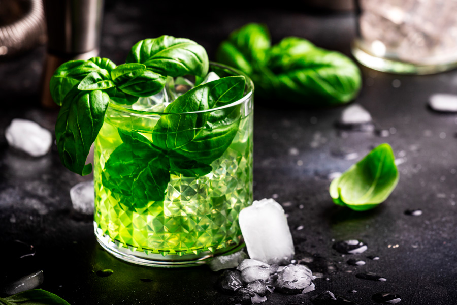 Immagine d'atmosfera: bicchiere pieno di liquore al basilico disposto con foglie di basilico e cubetti di ghiaccio su uno sfondo scuro