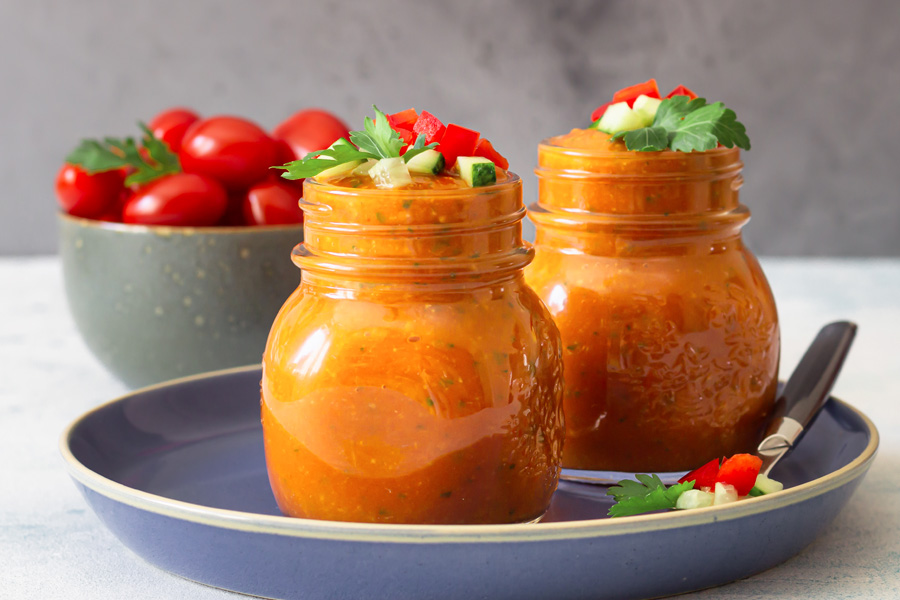 Ricetta del gazpacho Immagine d'atmosfera: due barattoli con tappo a vite pieni di gazpacho rosso disposti su un piatto blu, sullo sfondo una ciotola di pomodori