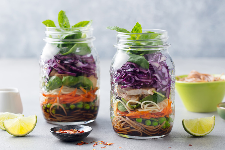 Immagine della ricetta Preparazione dei pasti: due grandi barattoli con tappo a vite pieni di pasta, carne e verdure crude, disposti con lime e peperoncino