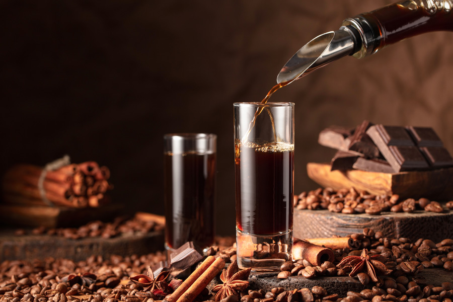 Ricetta del liquore al cioccolato Immagine della ricetta del liquore al cioccolato: il liquore viene versato da una bottiglia in un bicchierino, sullo sfondo c'è un altro bicchiere, disposto con cioccolato, chicchi di caffè e spezie